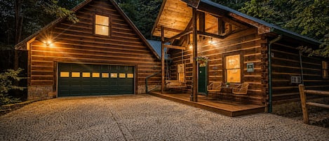 A log cabin with a stone-paved driveway, a green garage door, and a porch with outdoor seating.