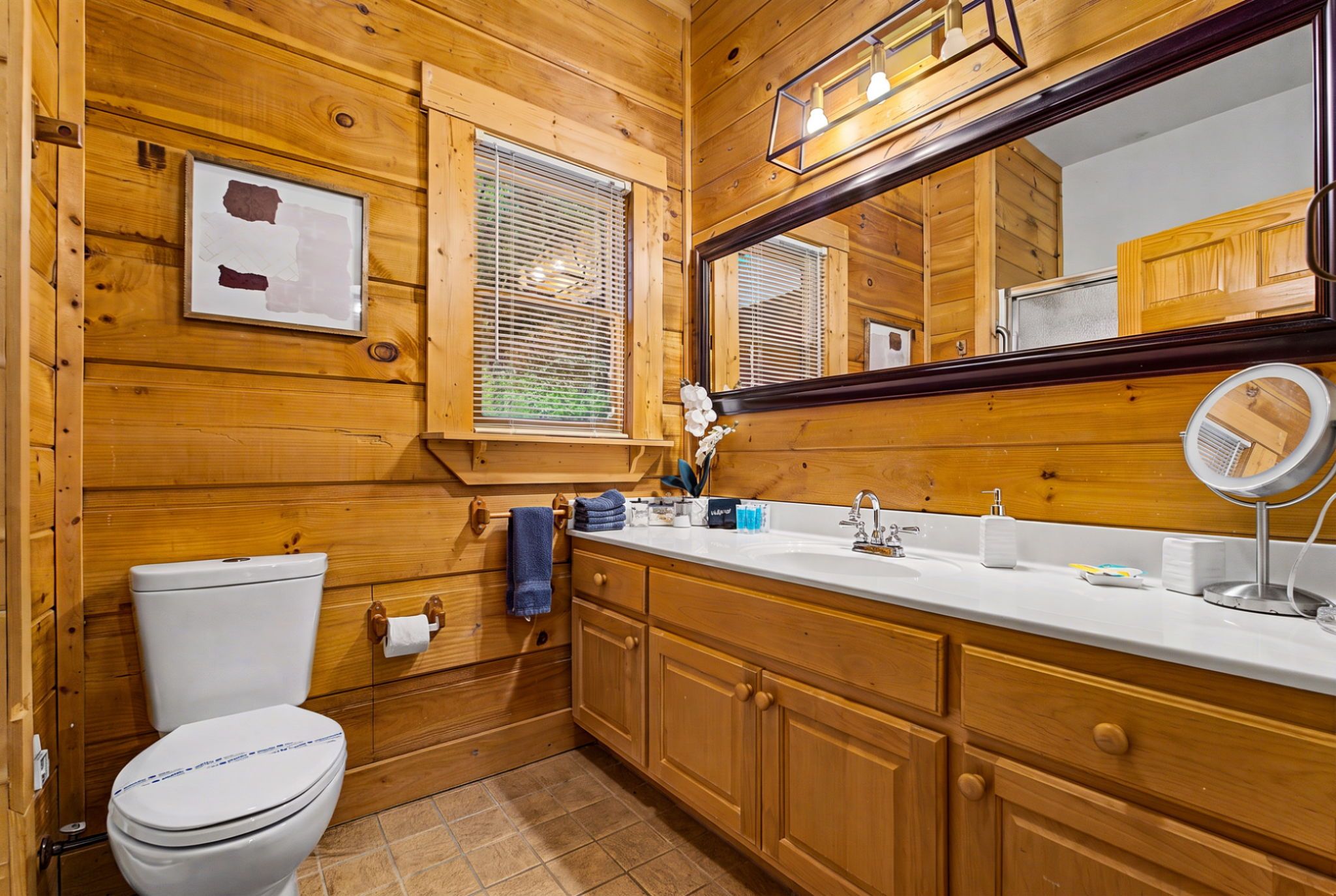 A wooden cabin-style bathroom with a toilet, sink, mirror, and window with blinds.