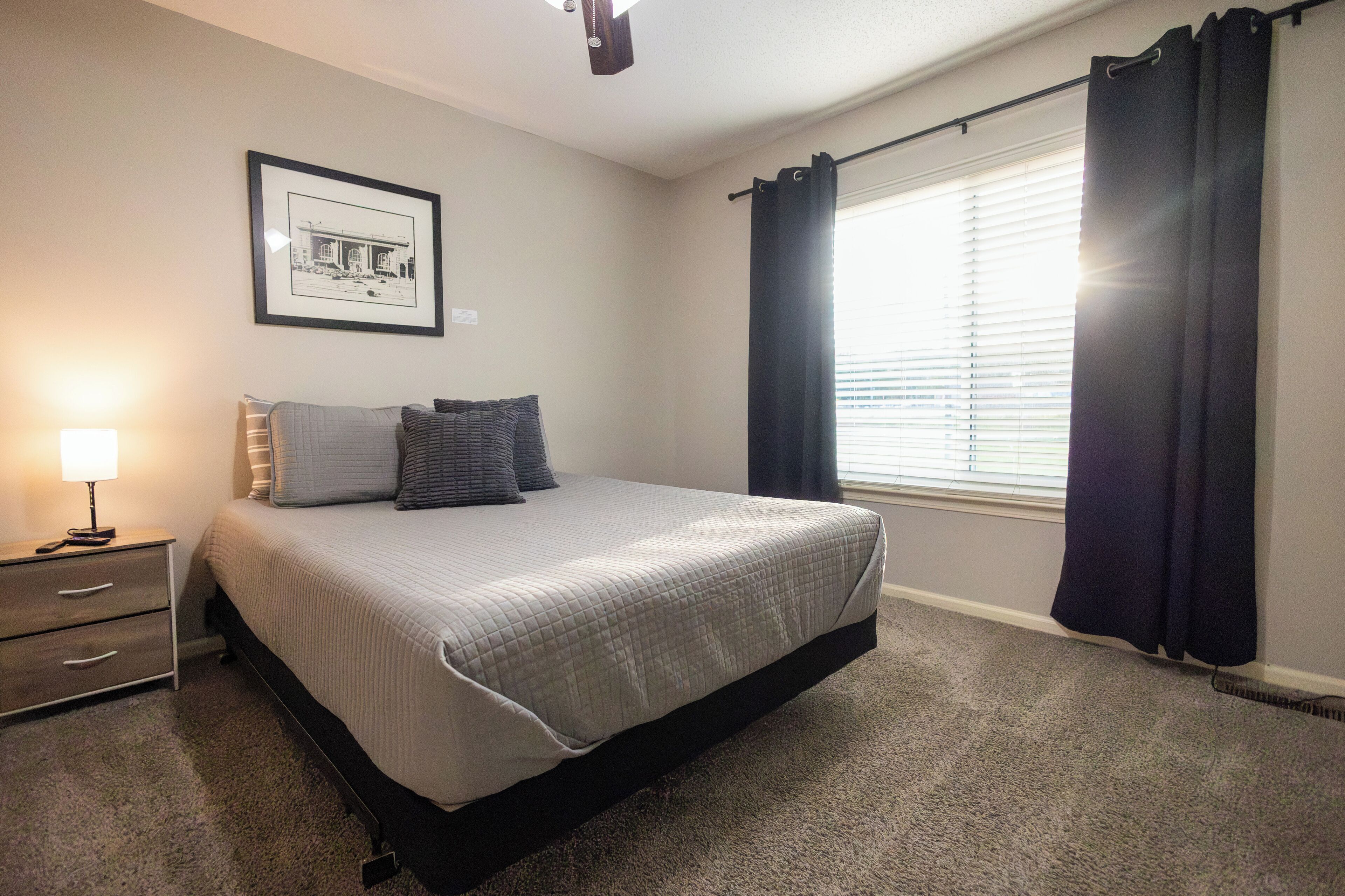 A bedroom with a bed, a nightstand, a lamp, a framed picture on the wall, and a window with curtains.