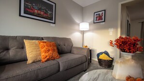 A living room with a grey sofa, orange and white cushions, a floor lamp, a vase with red flowers, and a framed picture on the wall. - Full clean 3-bedroom house close to stadiums and pet friendly! (Grandview)