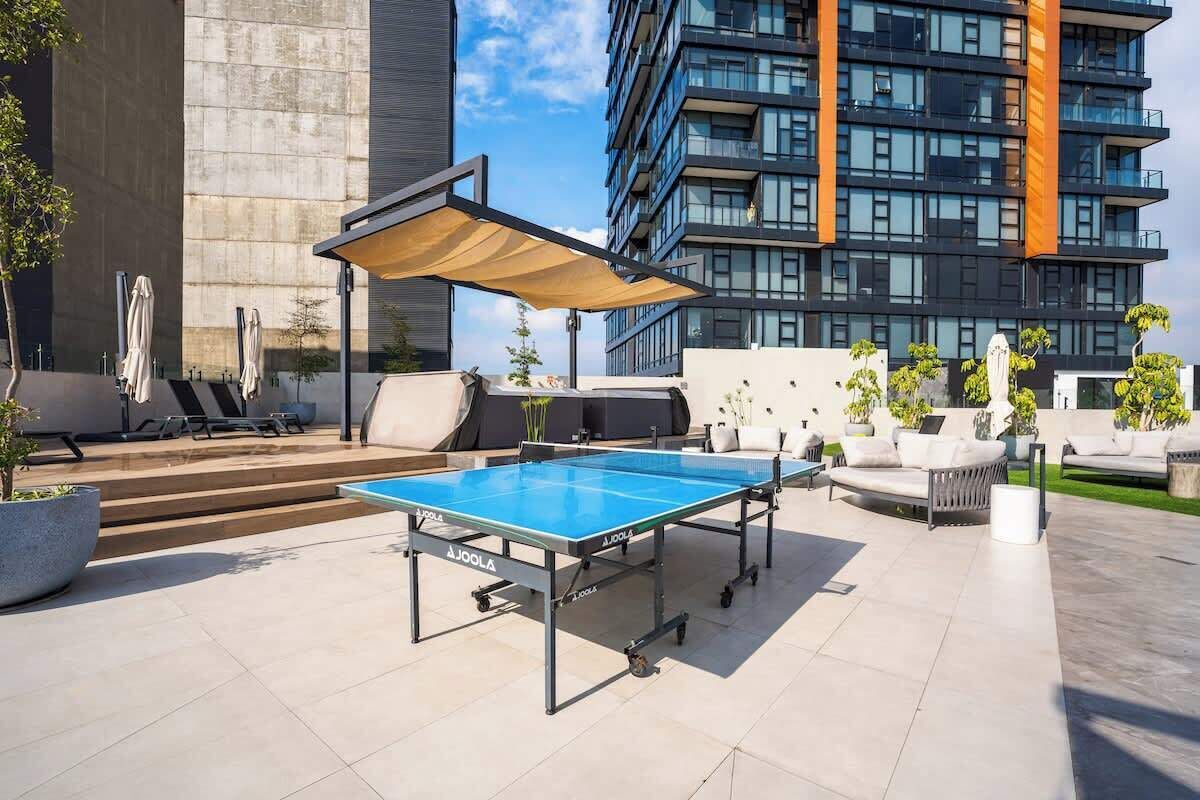 Mesa de ping pong al aire libre con toldo, rodeada de sillas de descanso y plantas en macetas, con un edificio moderno de fondo.