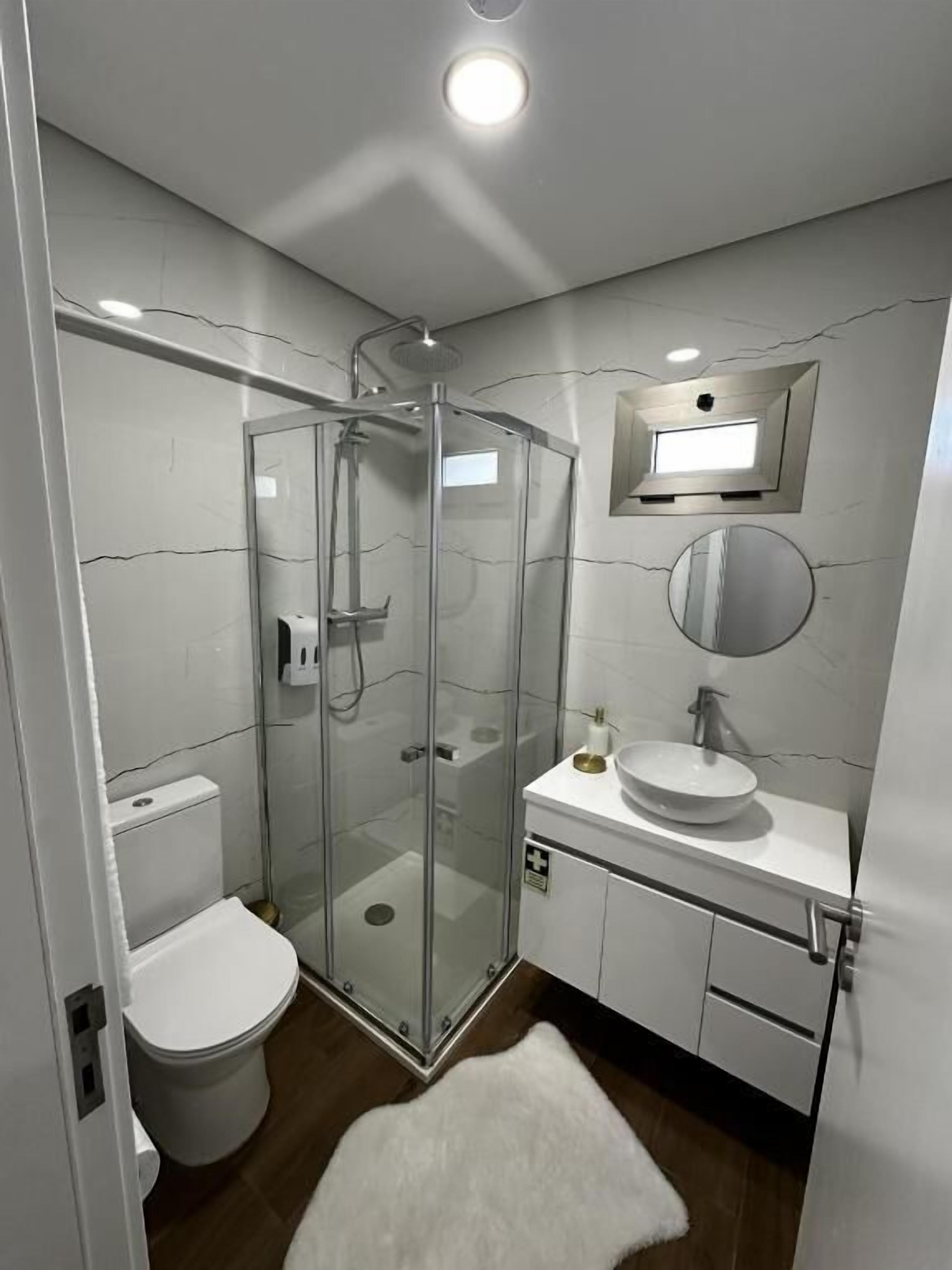 A modern bathroom with a glass-enclosed shower, a toilet, a sink with a mirror, and a white rug on the floor.