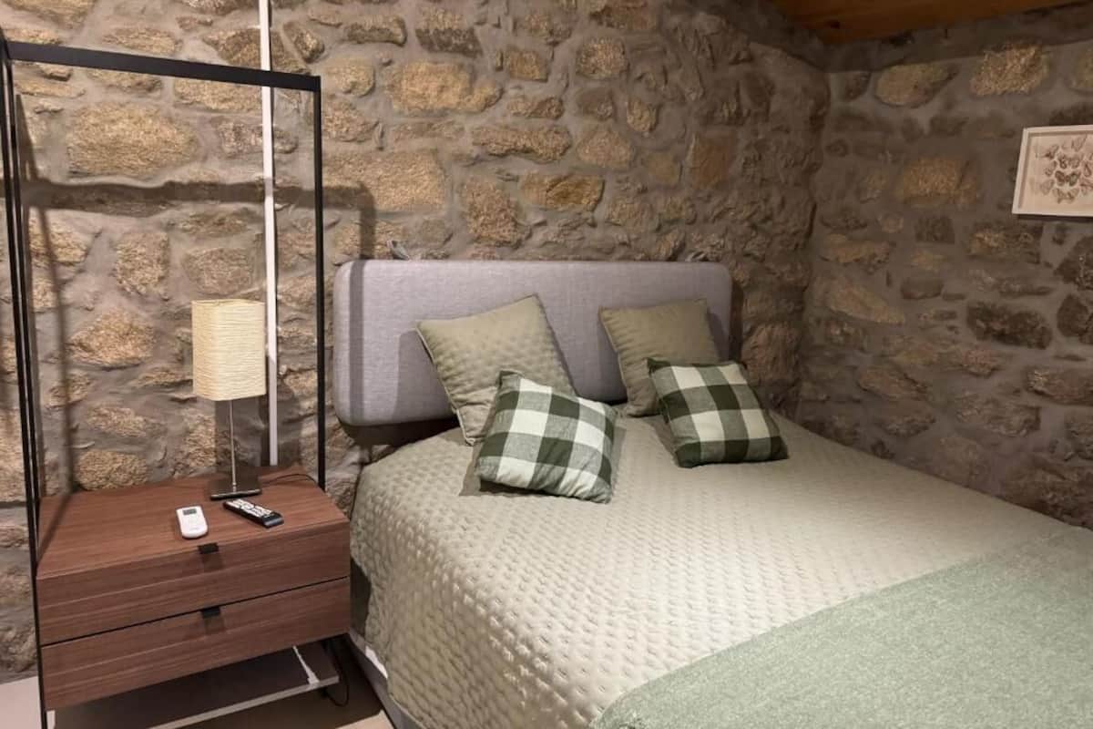 Une chambre avec un mur en pierre, un lit recouvert d’une courtepointe et agrémenté d’oreillers à carreaux, une table de chevet avec une lampe et une télécommande.