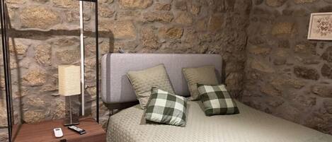 A bedroom with a stone wall, a bed with a quilt and checkered pillows, a bedside table with a lamp, and a remote control.