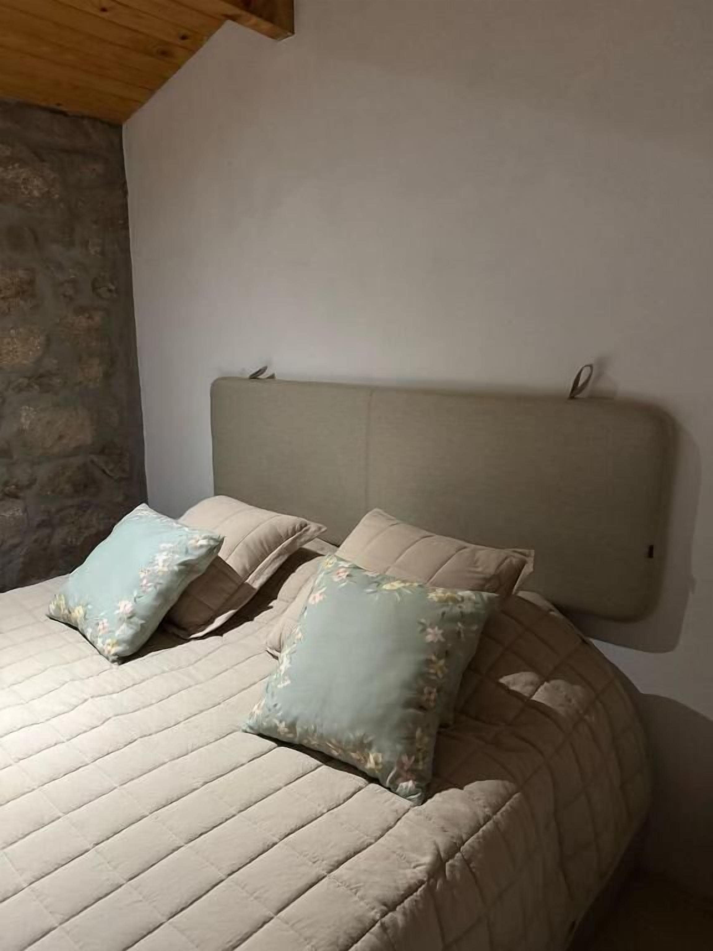 Una cama con cabecera acolchada, dos almohadas y una pared de piedra.