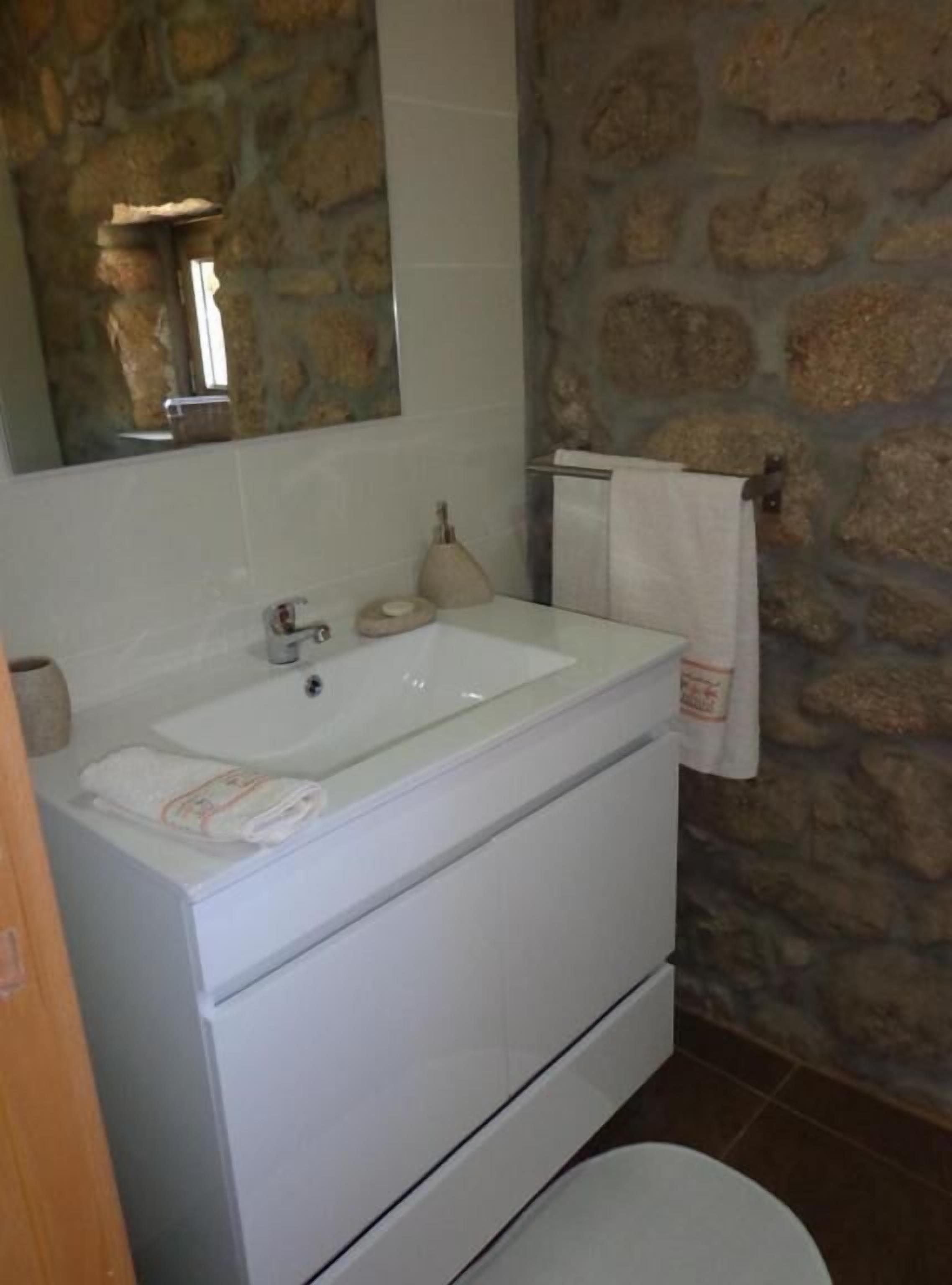 Un baño con pared de piedra, un lavabo blanco, un espejo y una toalla colgada en un perchero.