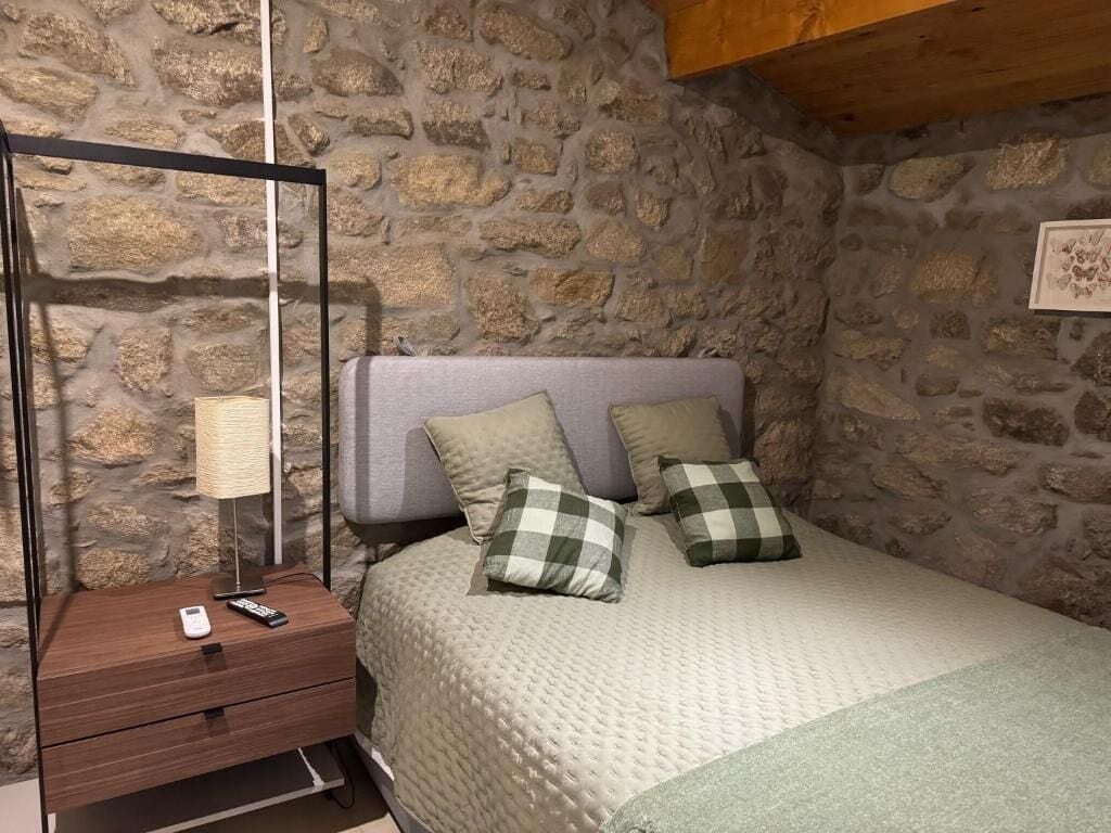 A bedroom with a stone wall, a bed with a grey quilt, two checkered pillows, a bedside table with a lamp, and a remote control.