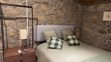 A bedroom with a stone wall, a bed with a grey quilt, two checkered pillows, a bedside table with a lamp, and a remote control.