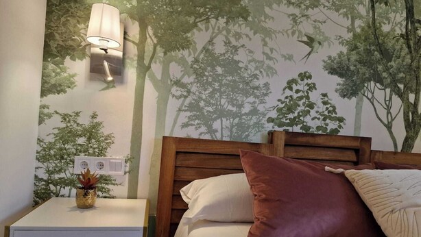 A hotel room with a bed, a nightstand with a lamp, and a wall mural of trees. - Mirador de Picos Apartment (Pido)