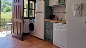 A compact kitchen with a washing machine, a refrigerator, and a dishwasher. A wooden door leads to an outdoor area with a view of greenery. - Mirador de Picos Apartment (Pido)