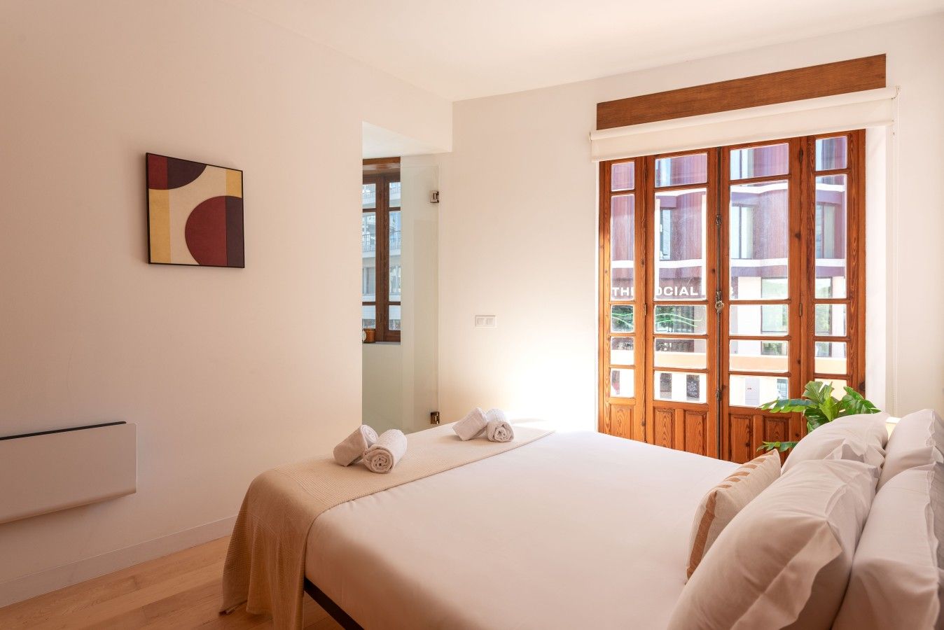 Quarto de hotel com cama, janela com persianas de madeira, um quadro na parede e um vaso de planta.