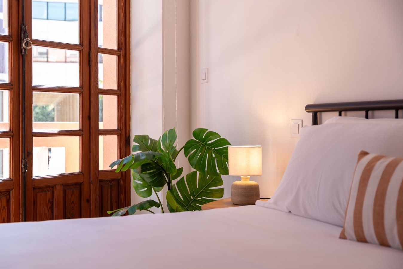 Une chambre d’hôtel avec un lit, une lampe de chevet, une plante en pot et une fenêtre avec des cadres en bois.