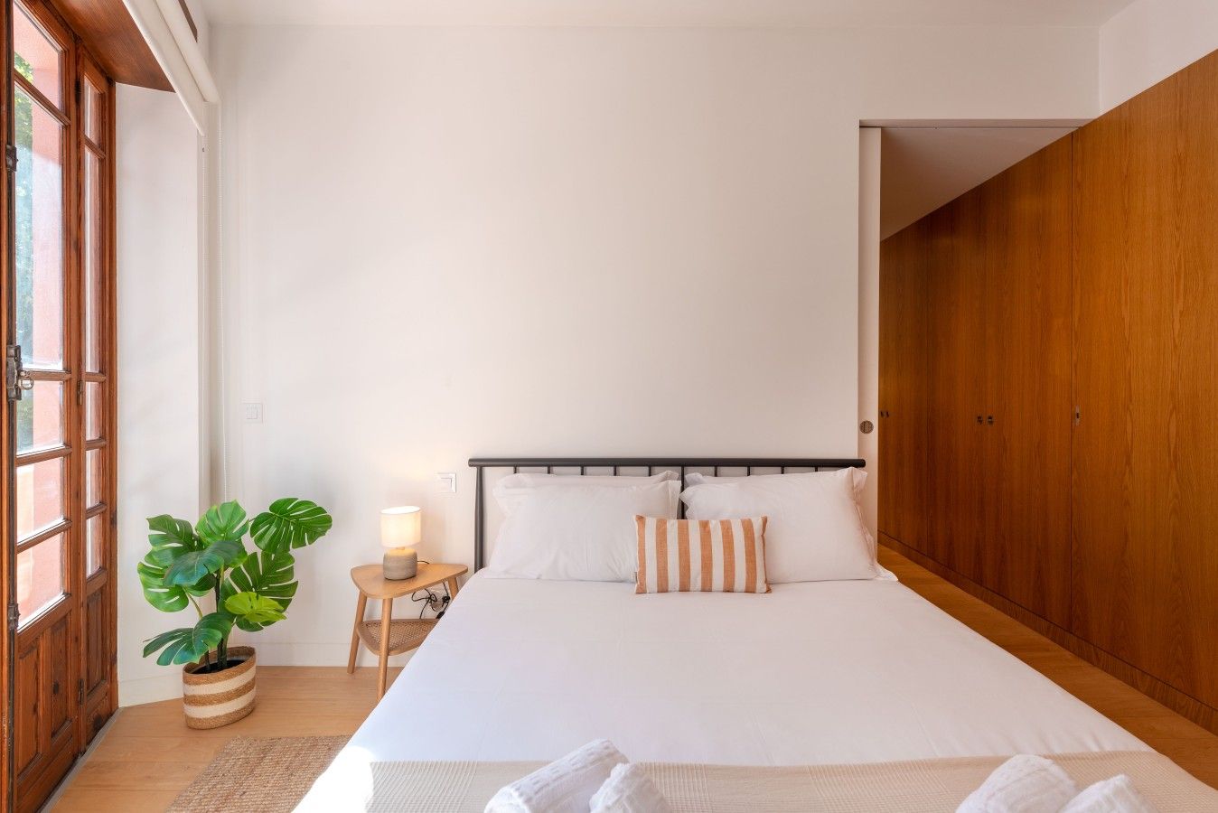 A bedroom with a bed, bedside table, lamp, potted plant, and wooden wardrobe.