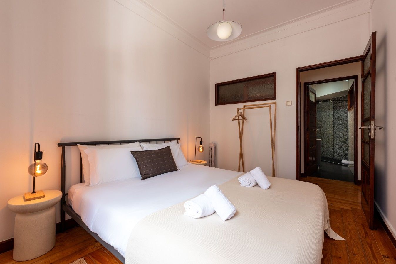 A neatly made bed with white linens and a brown pillow, a bedside table with a lamp, and a wooden floor.