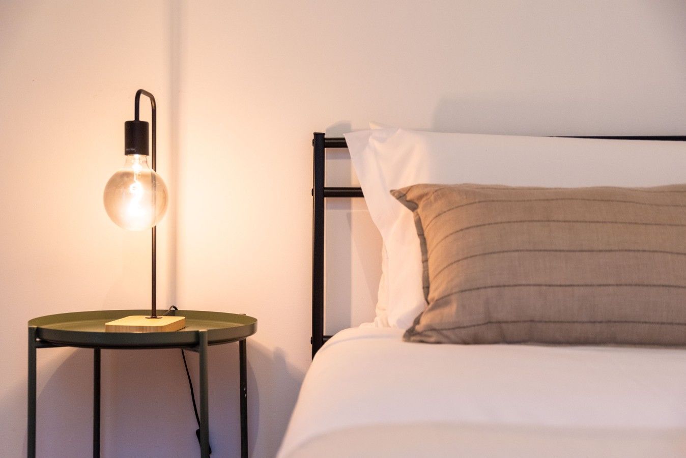 Ein Bett mit weißer Bettwäsche und einem einzelnen braunen Kopfkissen, ein runder Tisch mit einer schwarzen Lampe und ein schwarzer Metallrahmen.