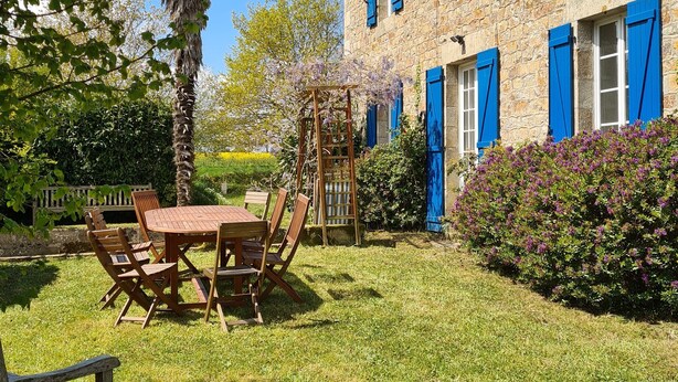 Een stenen gebouw met blauwe luiken, een houten tafel met stoelen, en een tuin met een palmboom en paarse bloemen.