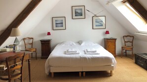 Een slaapkamer met een schuine plafond, een bed met witte lakens, twee stoelen, een houten bureau en een kleine tafel met een lamp.