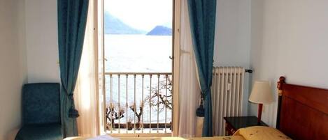 A hotel room with a bed, a chair, a nightstand, a lamp, and a view of a body of water and mountains.