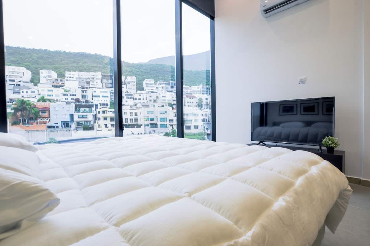 Un dormitorio con una cama grande, televisor y vistas a un paisaje urbano a través de amplias ventanas.