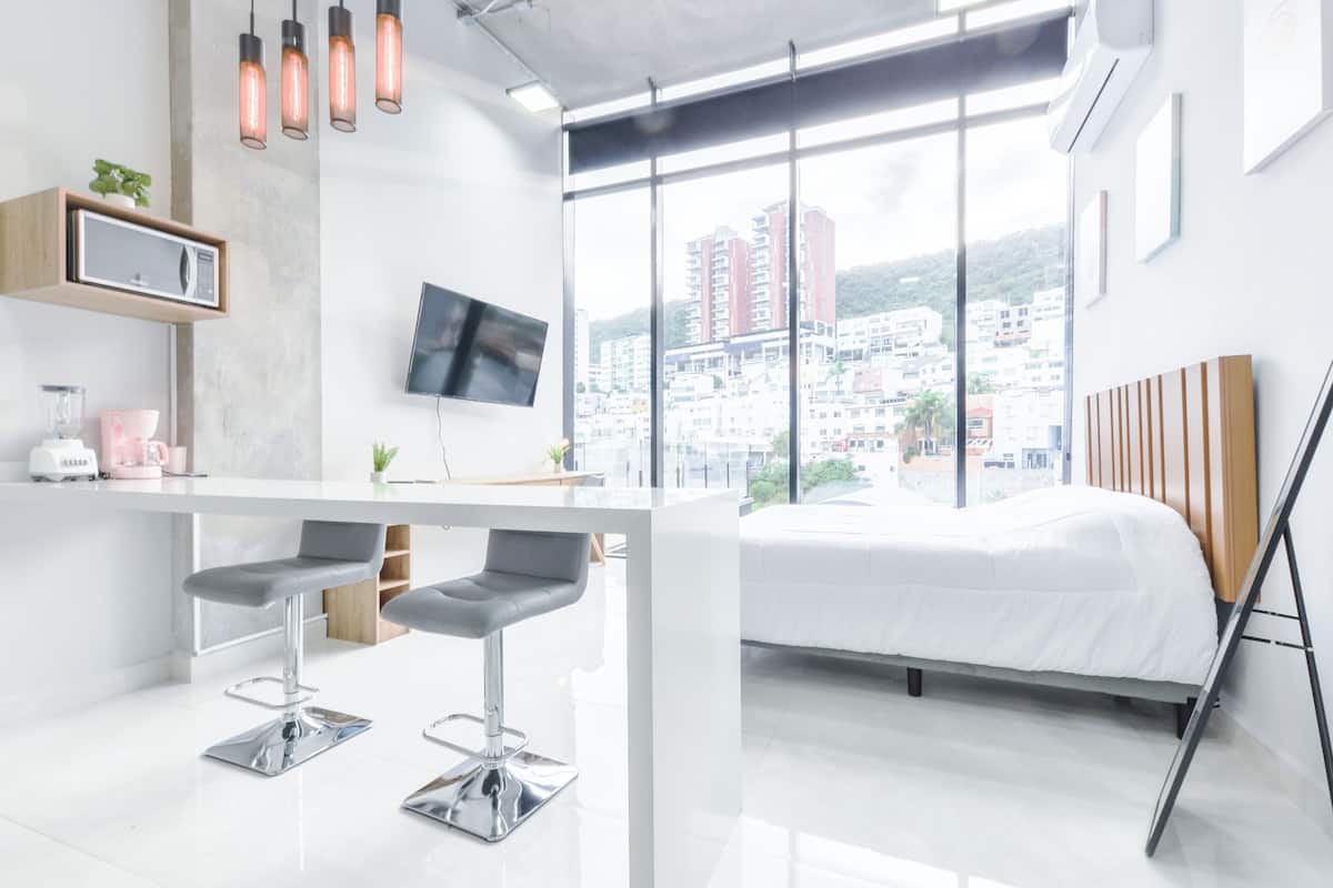 Una habitación de hotel moderna con un ventanal grande, una cama, un escritorio con dos taburetes altos y un televisor de pantalla plana, con vistas a la ciudad.