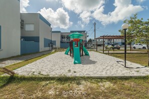 Um parquinho com escorrega e estruturas de escalada, uma área de estar coberta e um edifício com varandas ao fundo.
