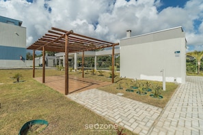 Um edifício moderno com pérgula de madeira, caminhos pavimentados e pequenas áreas ajardinadas.