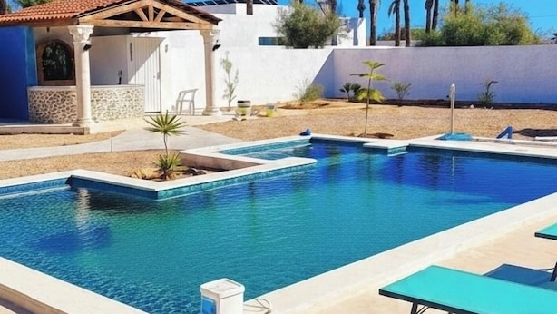 A swimming pool with lounge chairs, a covered seating area, and palm trees in the background. - Villa bleu azur 💙 (Djerba)