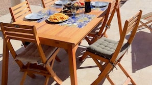 An outdoor dining area with a wooden table and chairs, a small building with a red-tiled roof, and a pool in the background. - Villa bleu azur 💙 (Djerba)