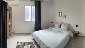 A bedroom with a bed, a nightstand, a window, and a shelf with decorative items. - Villa bleu azur 💙 (Djerba)