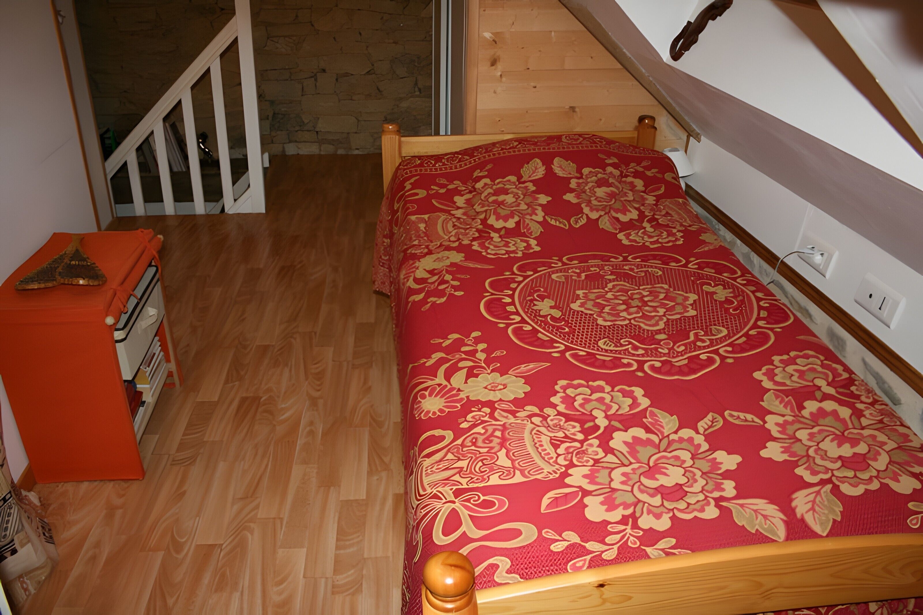 Una habitación pequeña con una cama de madera, una colcha roja con estampado, un aparador de madera y una escalera que conduce a un nivel superior.