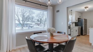 A modern kitchen with stainless steel appliances, a dining area with a wooden table and four chairs, and a large window with a view of a snowy landscape. - Remodeled Home w/ Garage, Workspace & Fenced Yard (Racine)