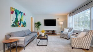 A modern living room with a grey sofa, patterned armchairs, a coffee table, and a flat-screen TV on the wall. - Remodeled Home w/ Garage, Workspace & Fenced Yard (Racine)