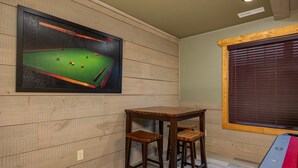A pool table with stools, a framed billiards cueing poster, and a window with blinds. - Mountain High (Gatlinburg)
