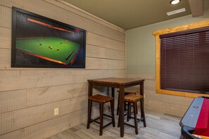 A pool table with stools, a framed billiards cueing poster, and a window with blinds. - Mountain High (Gatlinburg)