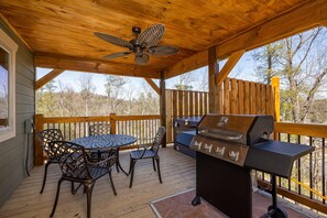 An outdoor deck with a wooden ceiling, a ceiling fan, a grill, and metal chairs. - Mountain High (Gatlinburg)