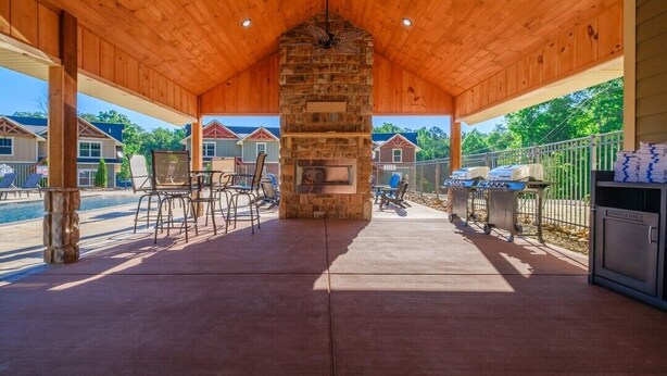 A covered patio with a stone fireplace, outdoor seating, and a pool in the background. - Mountain High (Gatlinburg)