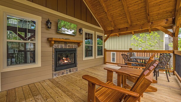 A wooden deck with a fireplace, outdoor seating, and large windows overlooking a forest. - Smoky Mountain Plunge (Gatlinburg)