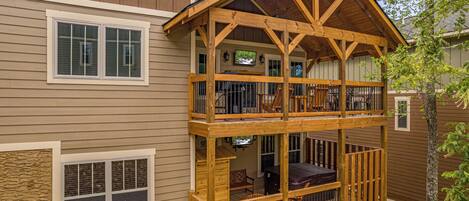 A two-story wooden deck with a balcony, a TV mounted on the wall, and a stone wall.
