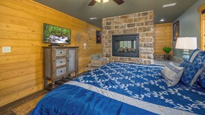A bedroom with a stone fireplace, wooden walls, a bed with blue bedding, a ceiling fan, and a television on a wooden cabinet. - Smoky Mountain Plunge (Gatlinburg)