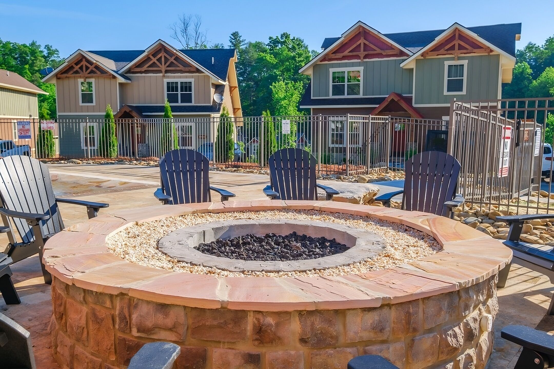 A circular stone fire pit surrounded by Adirondack chairs, with a paved patio area and a fence enclosing the space.