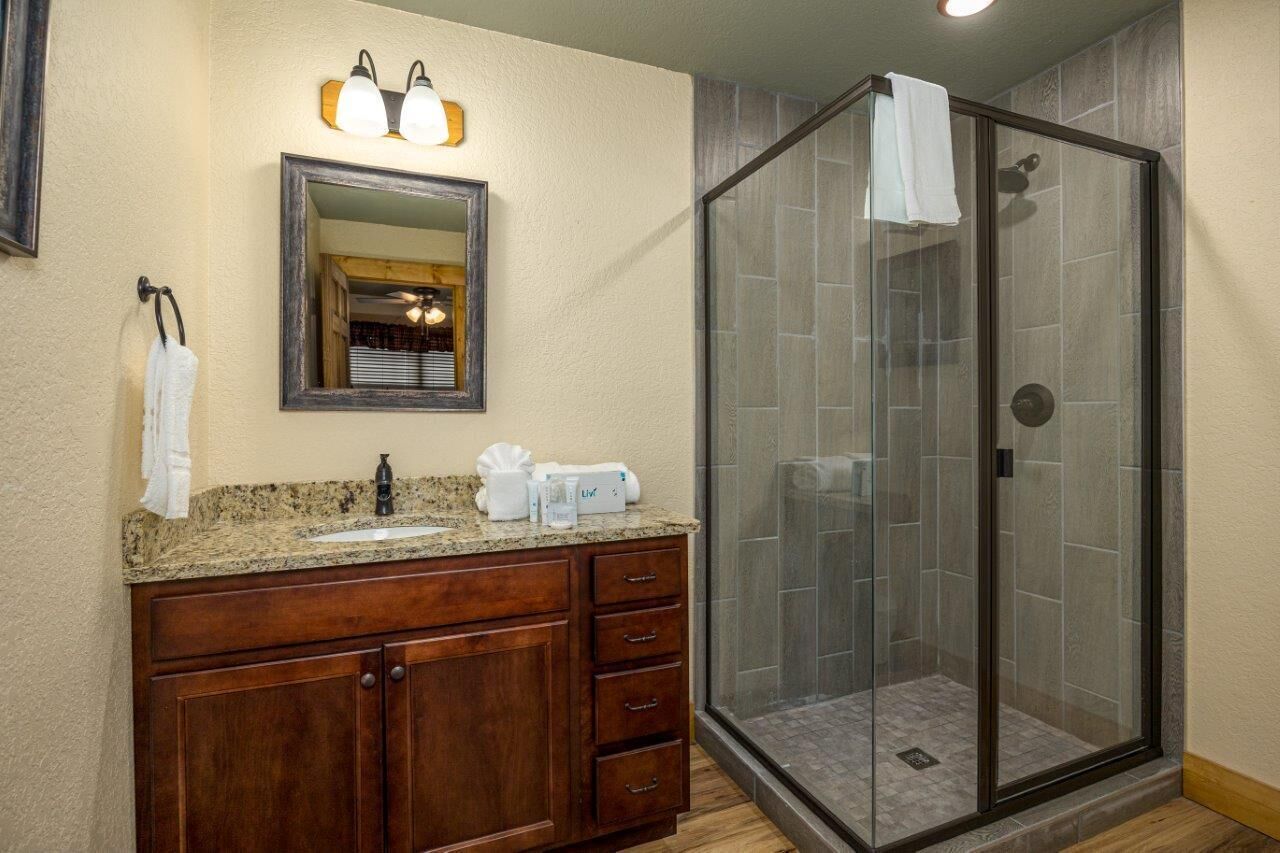 A bathroom with a granite countertop sink, wooden vanity, a mirror with wall-mounted lights, and a glass-enclosed shower.