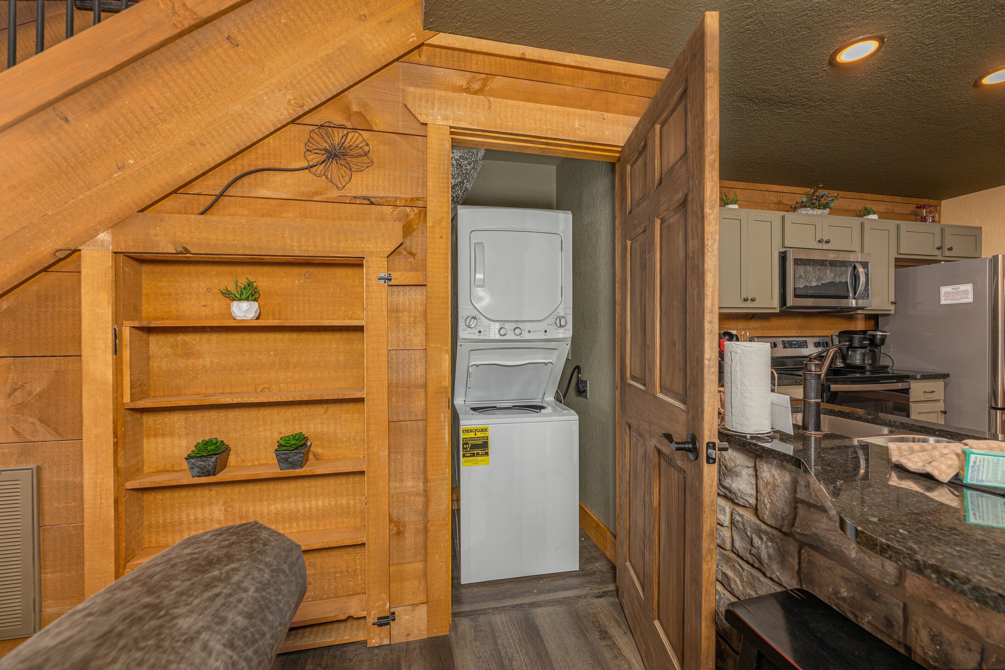 A compact kitchen with a built-in washer and dryer, wooden shelving with plants, and a stone accent wall.