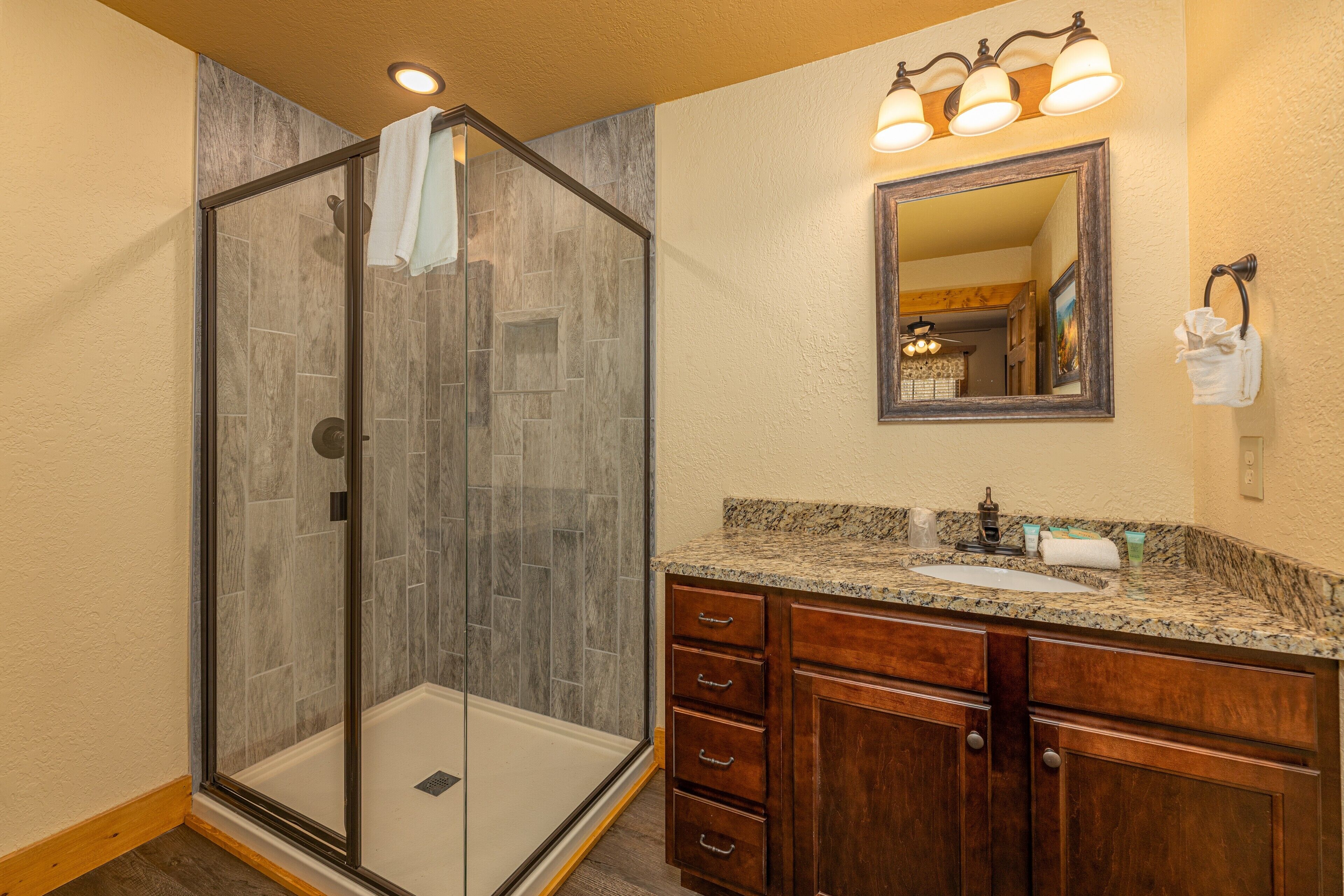 A bathroom with a glass-enclosed shower, a granite countertop, wooden cabinets, and a mirror with three lights.