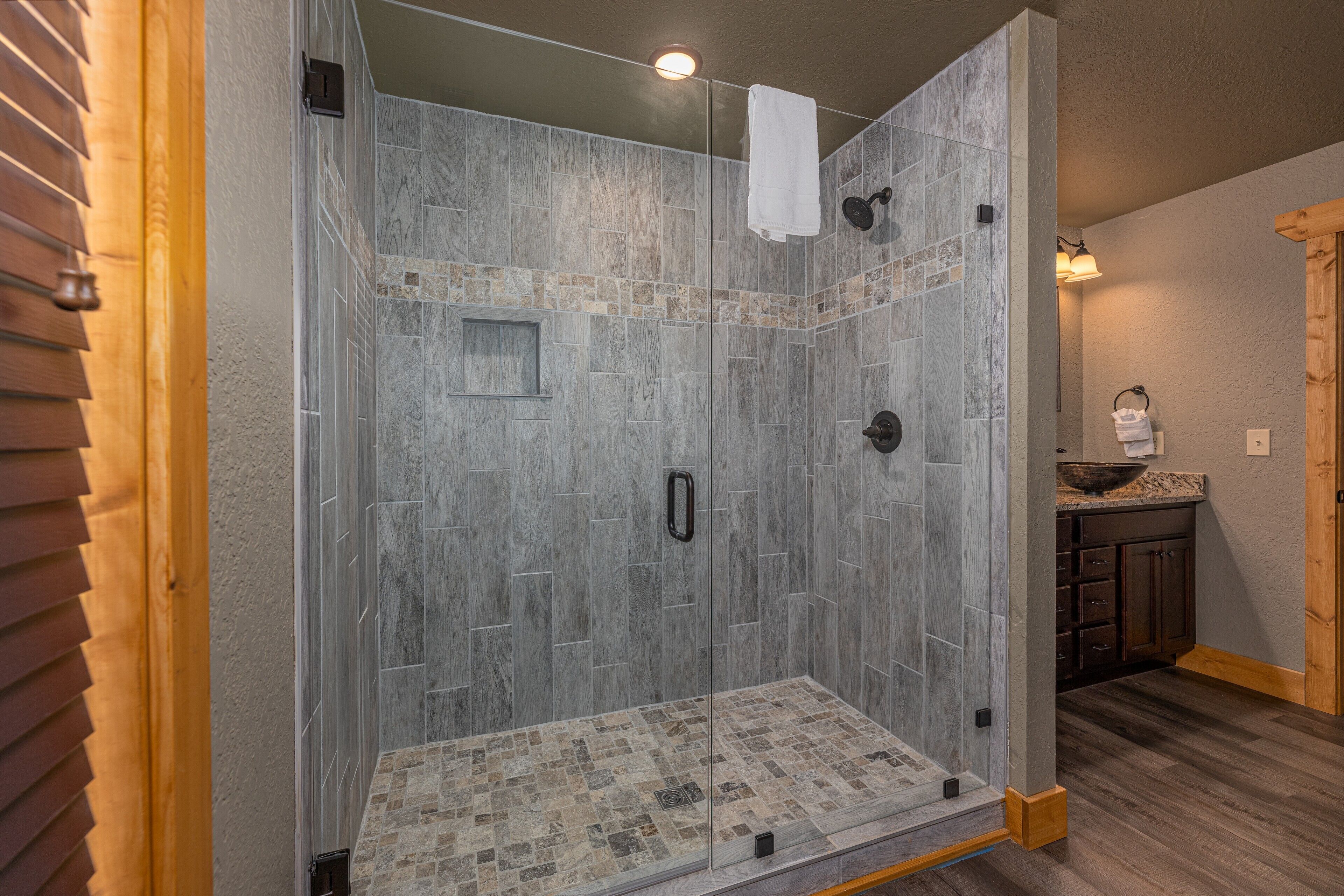 A walk-in shower with stone tiles, a glass door, and a wooden floor.
