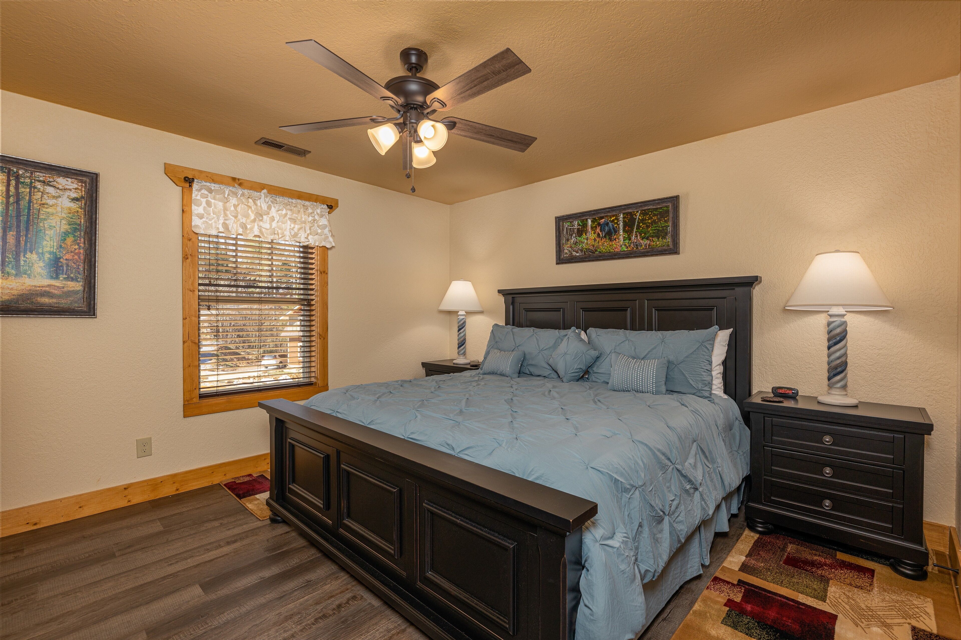 A bedroom with a large bed, a ceiling fan, two bedside tables with lamps, a window with blinds, and a painting on the wall.