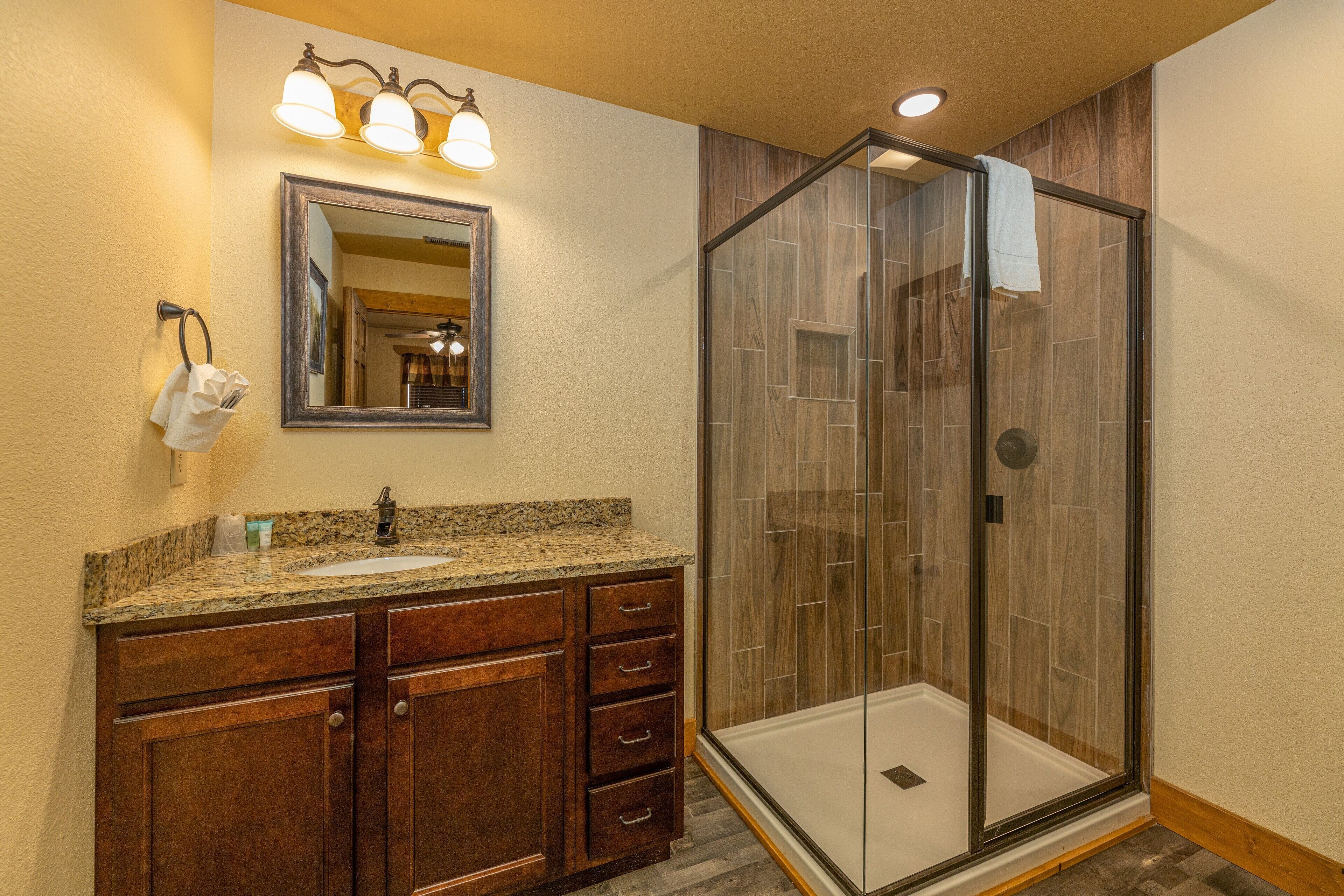 A bathroom with a granite countertop sink, wooden vanity, a mirror with lights, and a glass-enclosed shower.