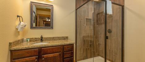 A bathroom with a granite countertop sink, wooden vanity, a mirror with lights, and a glass-enclosed shower.