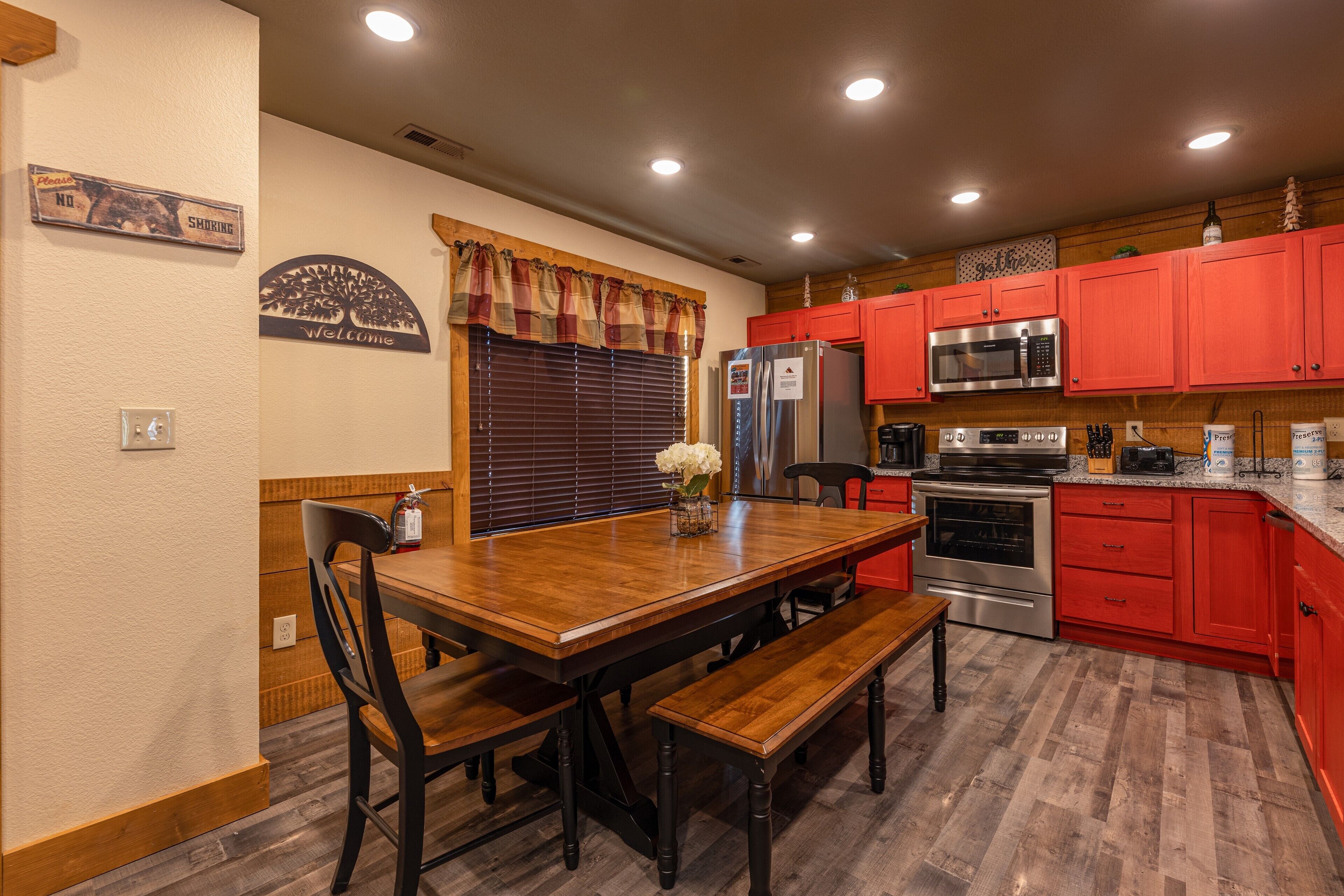 A kitchen with red cabinets, a wooden dining table with benches, a stainless steel refrigerator, and a microwave.
