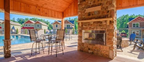 Un espacio al aire libre techado con chimenea de piedra, sillas junto a la piscina y vistas a casas vecinas.