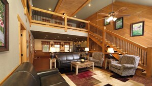 A spacious living area with a black leather sofa, patterned armchairs, and a wooden coffee table. A staircase with a wooden railing leads to an upper level. - Head Over Hills (Gatlinburg)